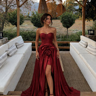 Woman wearing a ruby red couture evening gown with a structured corset bodice, oversized rose detail at the waist, high slit, and dramatic train.