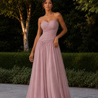 Woman wearing the Rosalie Evening Dress, a blush pink strapless gown with a draped sweetheart bodice and flowing floor-length skirt in a garden setting.