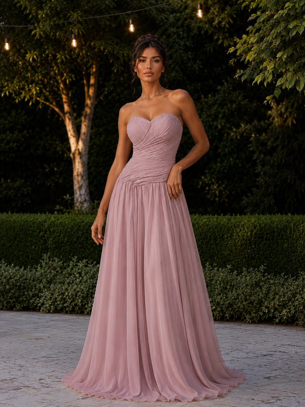 Woman wearing the Rosalie Evening Dress, a blush pink strapless gown with a draped sweetheart bodice and flowing floor-length skirt in a garden setting.
